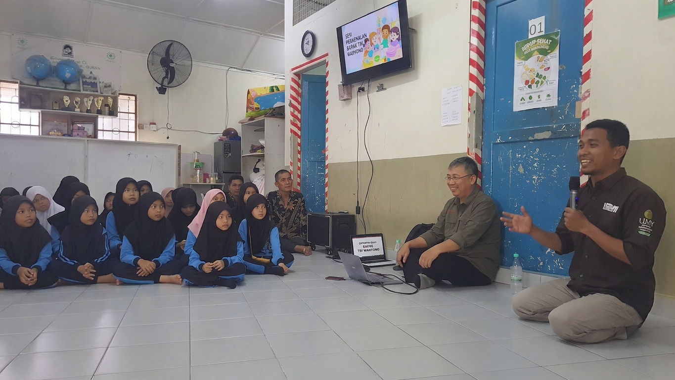 Children in hijabs sitting on floor listening to two men speaking with a microphone indoors